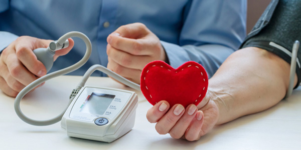 Person hält ein rotes, herzförmiges Objekt, während eine zweite Person den Blutdruck am Arm misst. Blutdruckmessgerät liegt auf dem Tisch, Manschette ist am Arm befestigt.